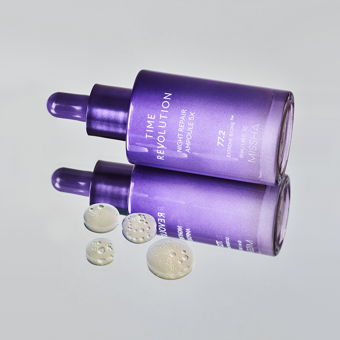 Two bottles of MISSHA Time Revolution Night Repair Ampoule 5X in gradient purple packaging lying on a reflective surface, surrounded by drops of the serum showing its smooth, hydrating texture with visible microbubbles.