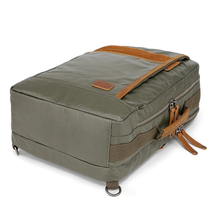 The TSD BRAND Madrone Coated Canvas Backpack in army green features brown leather accents, zippers, a top flap, side handles, metal rings, and a leather logo patch; shown lying flat to display these details.
