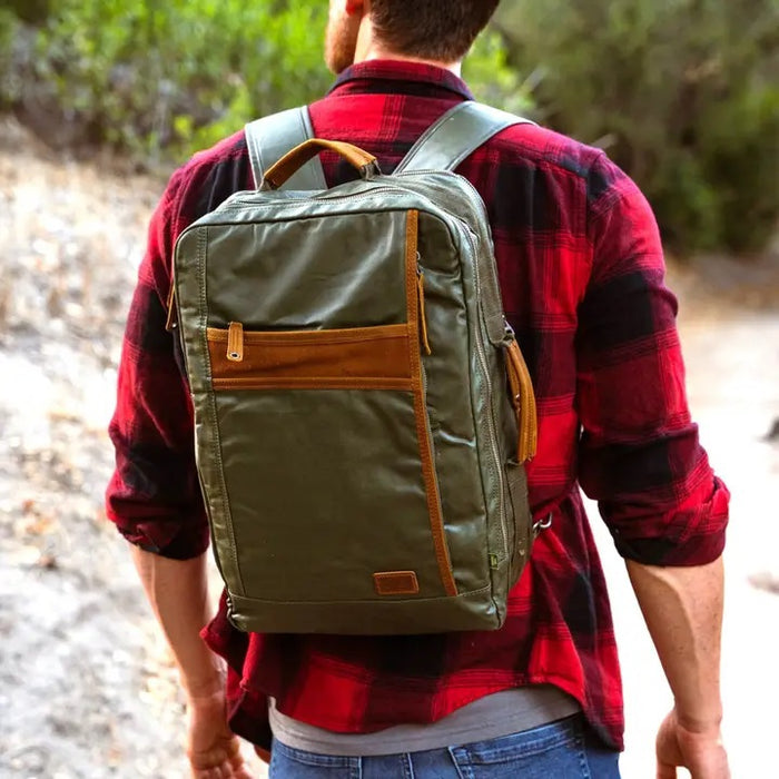 The TSD BRAND Madrone Coated Canvas Backpack in army green features brown leather accents, zippers, a top flap, side handles, metal rings, and a leather logo patch; shown lying flat to display these details.