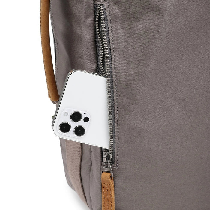 A close-up of the TSD BRAND - MADRONE Coated Canvas Backpack in Grey shows its brown leather accents and an unzipped front pocket with a black wallet partially visible inside.