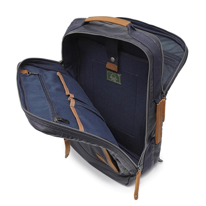The TSD BRAND - MADRONE COATED CANVAS BACKPACK IN NAVY features brown leather accents, a spacious main compartment, a padded laptop sleeve with snap closure, and multiple zippered pockets on the inside flap.
