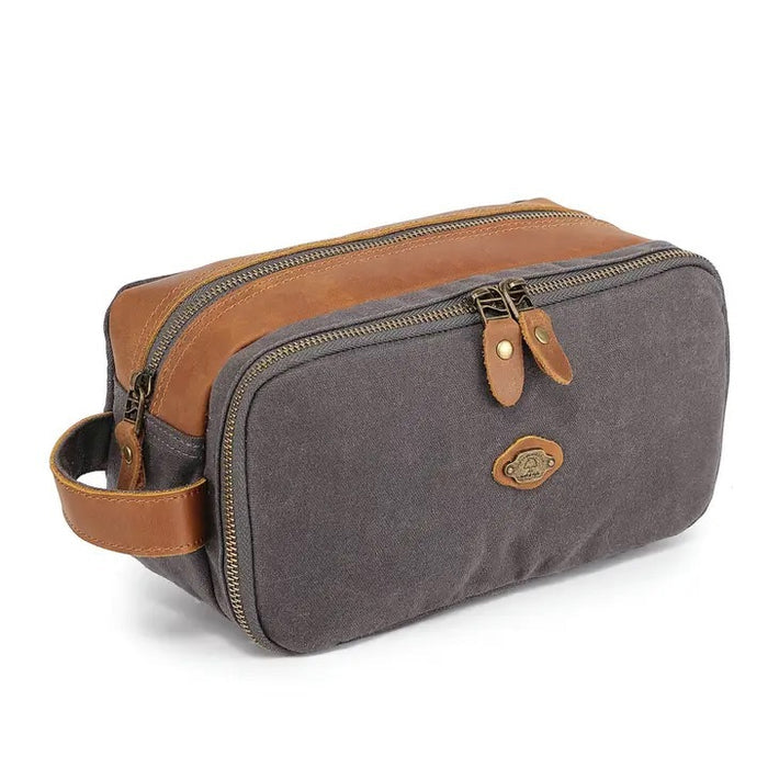 The TSD BRAND - Turtle Ridge Toiletry Bag in grey features waxed canvas, leather details, and a zip closure. It is open to show neatly organized travel essentials inside—bottles, a tube, a brush, and a white rectangular item.