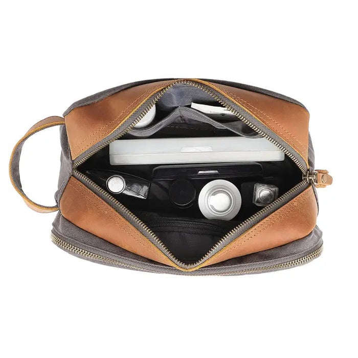 The TSD BRAND - Turtle Ridge Toiletry Bag in grey features waxed canvas, leather details, and a zip closure. It is open to show neatly organized travel essentials inside—bottles, a tube, a brush, and a white rectangular item.