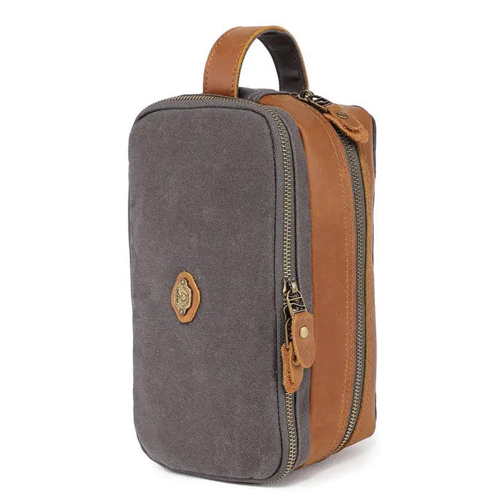 The TSD BRAND - Turtle Ridge Toiletry Bag in grey features waxed canvas, leather details, and a zip closure. It is open to show neatly organized travel essentials inside—bottles, a tube, a brush, and a white rectangular item.