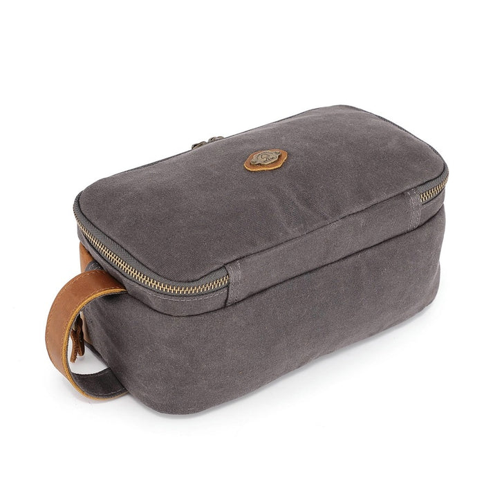 The TSD BRAND - Turtle Ridge Toiletry Bag in grey features waxed canvas, leather details, and a zip closure. It is open to show neatly organized travel essentials inside—bottles, a tube, a brush, and a white rectangular item.