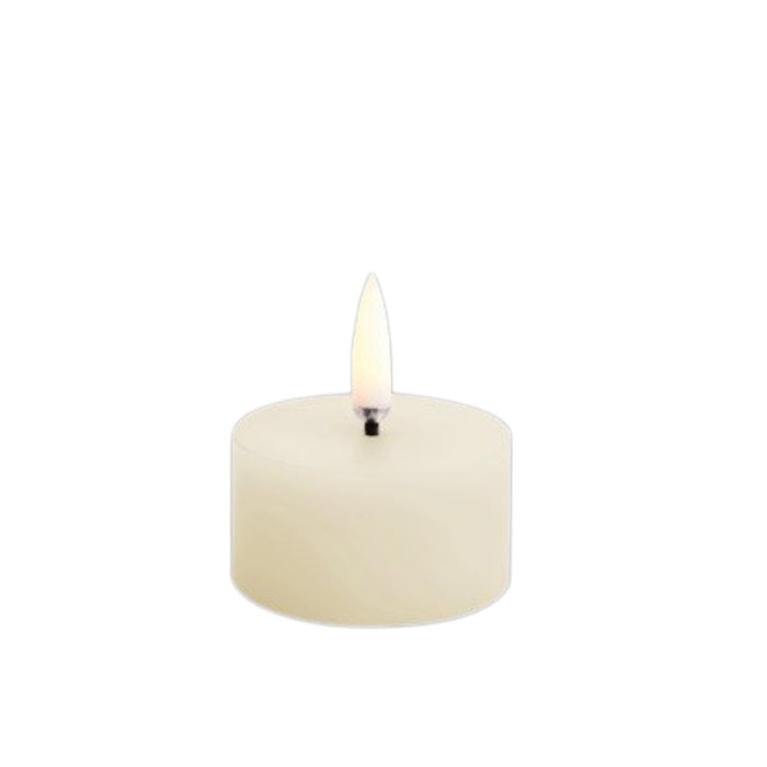 A lit PIFFANY UYUNI - IVORY PILLAR CANDLE 2X1.1 sits in a glass and stone holder on a wooden tray beside a small tealight candle and two gold rings, with a brown leather cushion in the background.