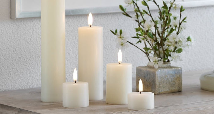 A lit PIFFANY UYUNI - IVORY PILLAR CANDLE 2X1.1 sits in a glass and stone holder on a wooden tray beside a small tealight candle and two gold rings, with a brown leather cushion in the background.