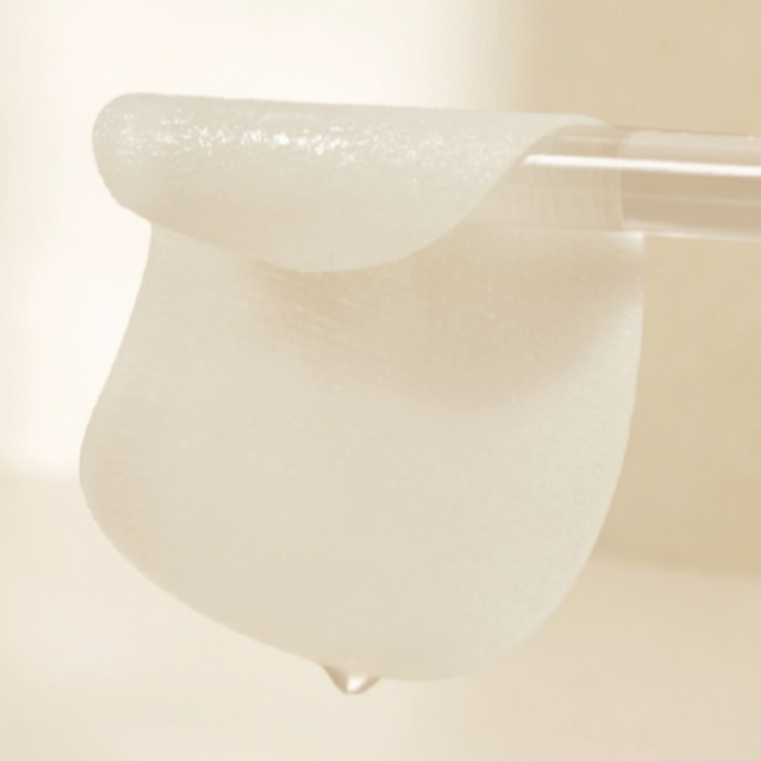A close-up of a moist skincare pad draped over a clear glass rod, with a droplet of essence falling from the pad, highlighting its hydration and texture.