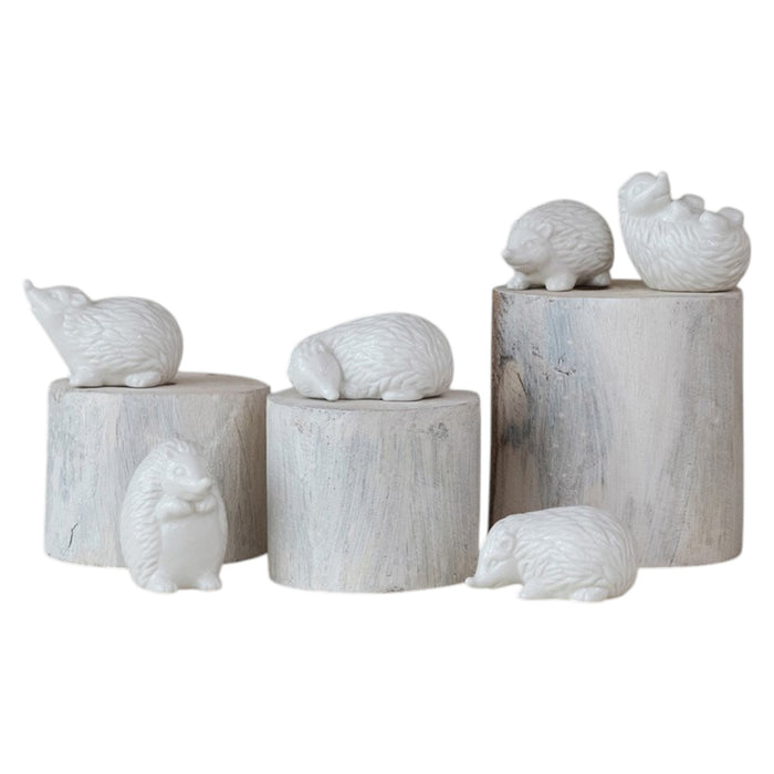 Six CREATIVE COOP WHITE STONEWARE HEDGEHOG figurines in different poses are arranged on and around three whitewashed cylindrical wooden pedestals, perfect for charming farmhouse decor. The background is plain white.