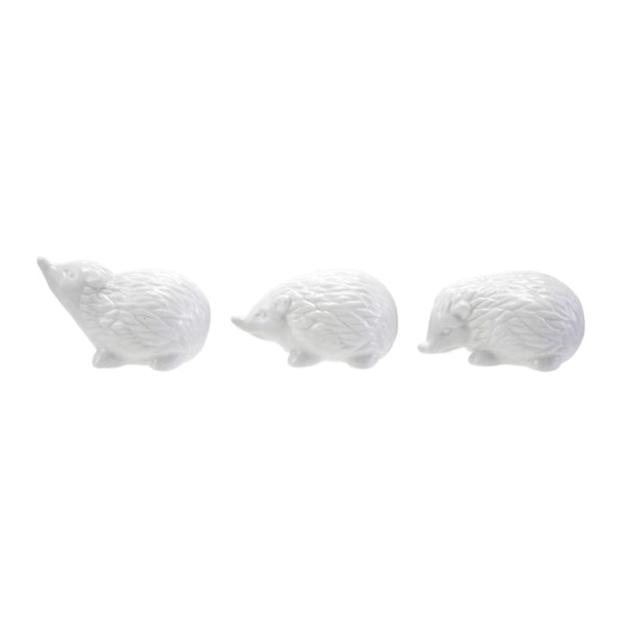 Six CREATIVE COOP WHITE STONEWARE HEDGEHOG figurines in different poses are arranged on and around three whitewashed cylindrical wooden pedestals, perfect for charming farmhouse decor. The background is plain white.