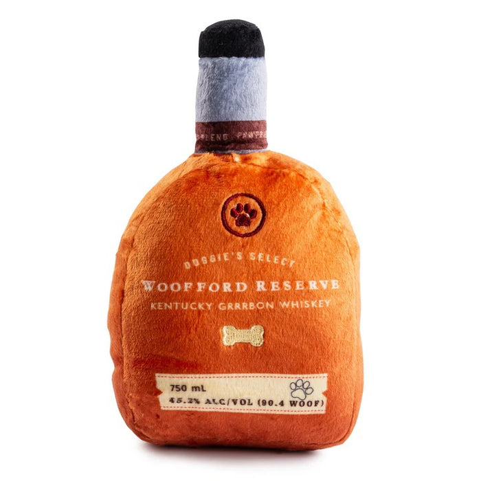 A golden retriever holds a HAUTE DIGGITY DOG WOOFFORD RESERVE BOURBON DOG TOY, a plush squeaky toy shaped like a whiskey bottle with "Woofford Reserve Kentucky" on the label. The dog's nose and eyes are visible, and the plush features a tag displaying volume and alcohol content.