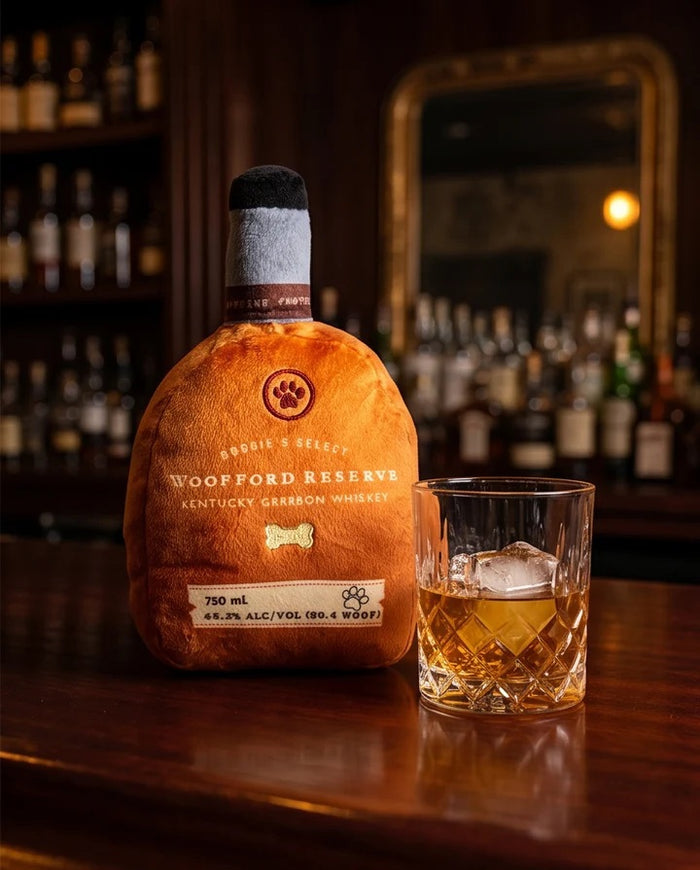 A golden retriever holds a HAUTE DIGGITY DOG WOOFFORD RESERVE BOURBON DOG TOY, a plush squeaky toy shaped like a whiskey bottle with "Woofford Reserve Kentucky" on the label. The dog's nose and eyes are visible, and the plush features a tag displaying volume and alcohol content.