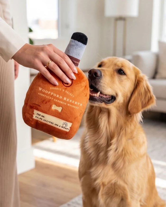 A golden retriever holds a HAUTE DIGGITY DOG WOOFFORD RESERVE BOURBON DOG TOY, a plush squeaky toy shaped like a whiskey bottle with "Woofford Reserve Kentucky" on the label. The dog's nose and eyes are visible, and the plush features a tag displaying volume and alcohol content.